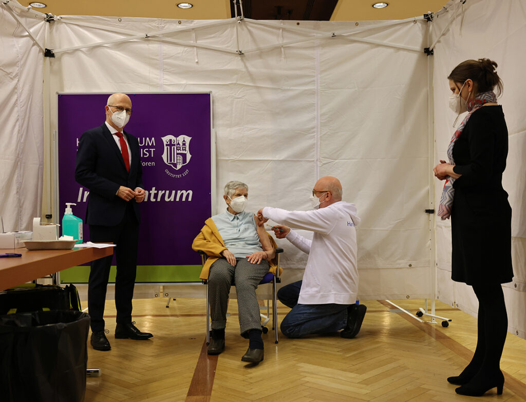 Hoffnung 2021: Erste Corona-Impfung in Hamburg. Am Sonntag nach Weihnachten wurden die ersten über 80-Jährigen gegen Corona geimpft. In Anwesenheit von Bürgermeister Peter Tschentscher und Sozialsenatorin Melanie Leonhard (r.) startete Dirk Heinrich (M.r.), ärztlicher Leiter des Impfzentrums Hamburg an den Messehallen, den Impfmarathon gegen die Pandemie. © picture alliance/dpa | Christian Charisius