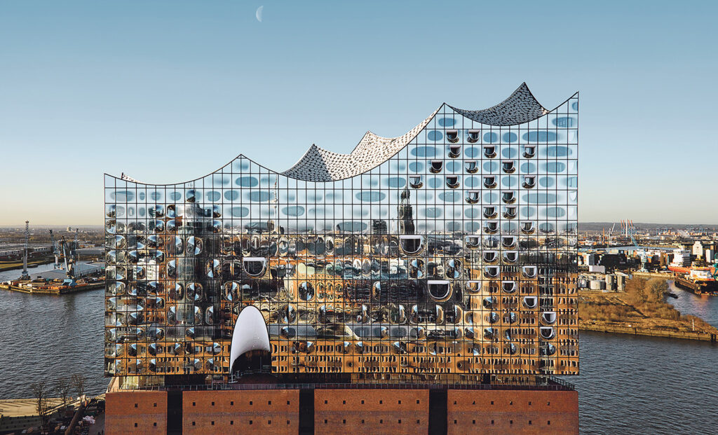 Elbphilharmonie, 2018: Das gläserne Jetztzeit-Wahrzeichen Elbphilharmonie kontert mit seiner gläsernen Fassade als Wellensymphonie die gnadenlose Sachlichkeit des einfachen roten Lagerhaus-Backsteinsockels des früheren Kaispeicher A. © Architekturbüro: Herzog & de Meuron, Basel, Schweiz; Foto: Maxim Schulz